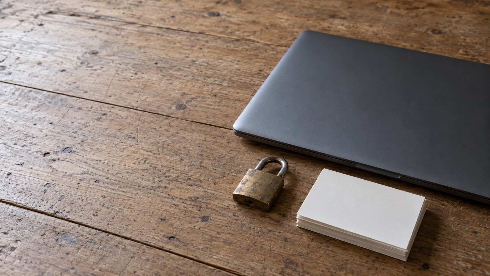 A closed laptop beside a small brass padlock and a stack of blank index cards on a wooden desk.