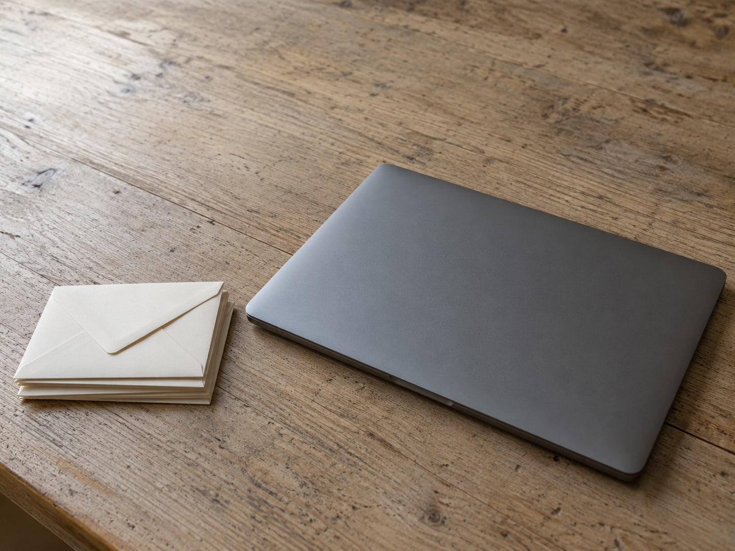 A closed laptop beside a small stack of unopened envelopes on a worn wooden desk.