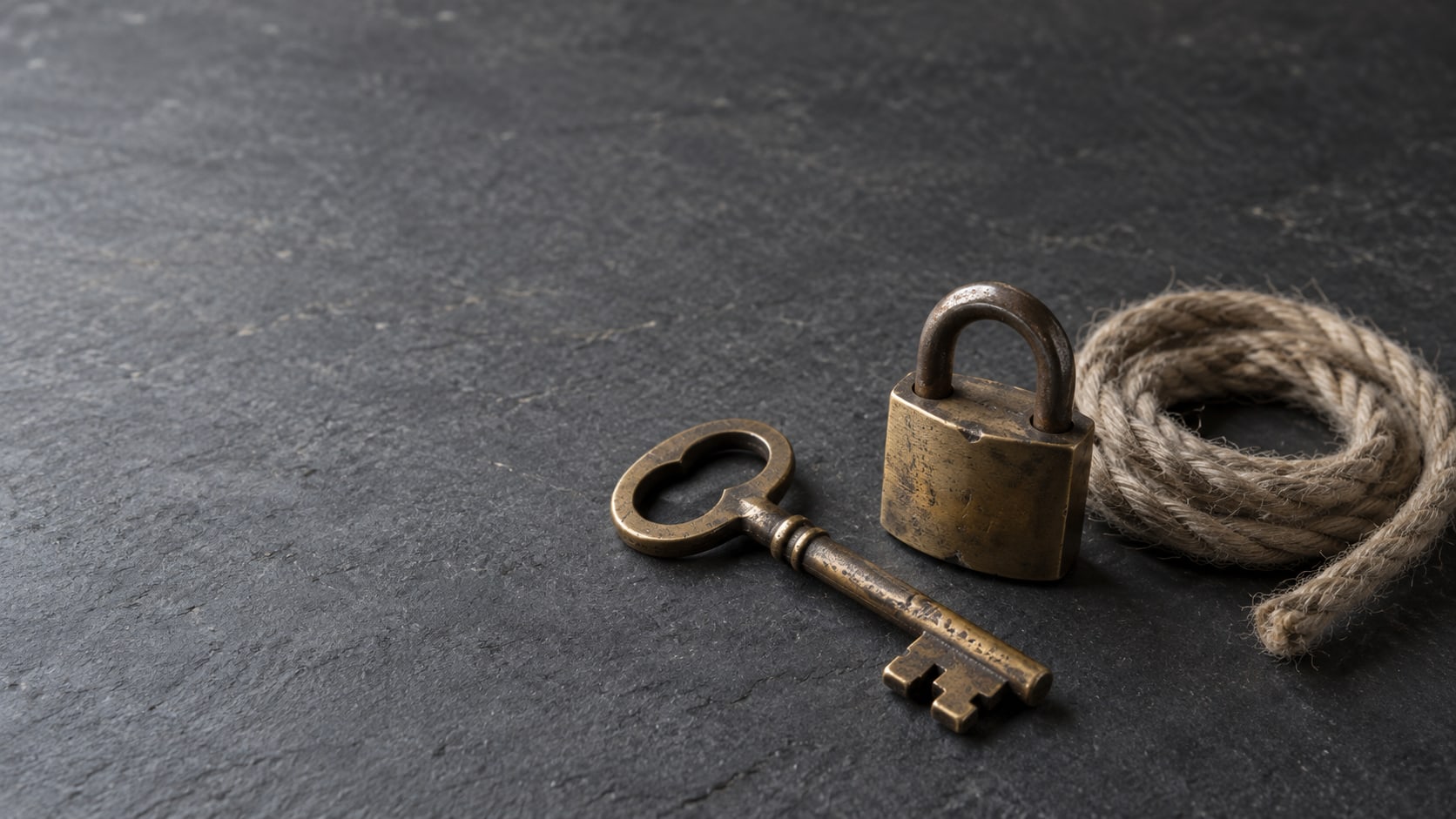 A weathered brass key and a small padlock on a plain slate surface, with a single coil of rope beside them.