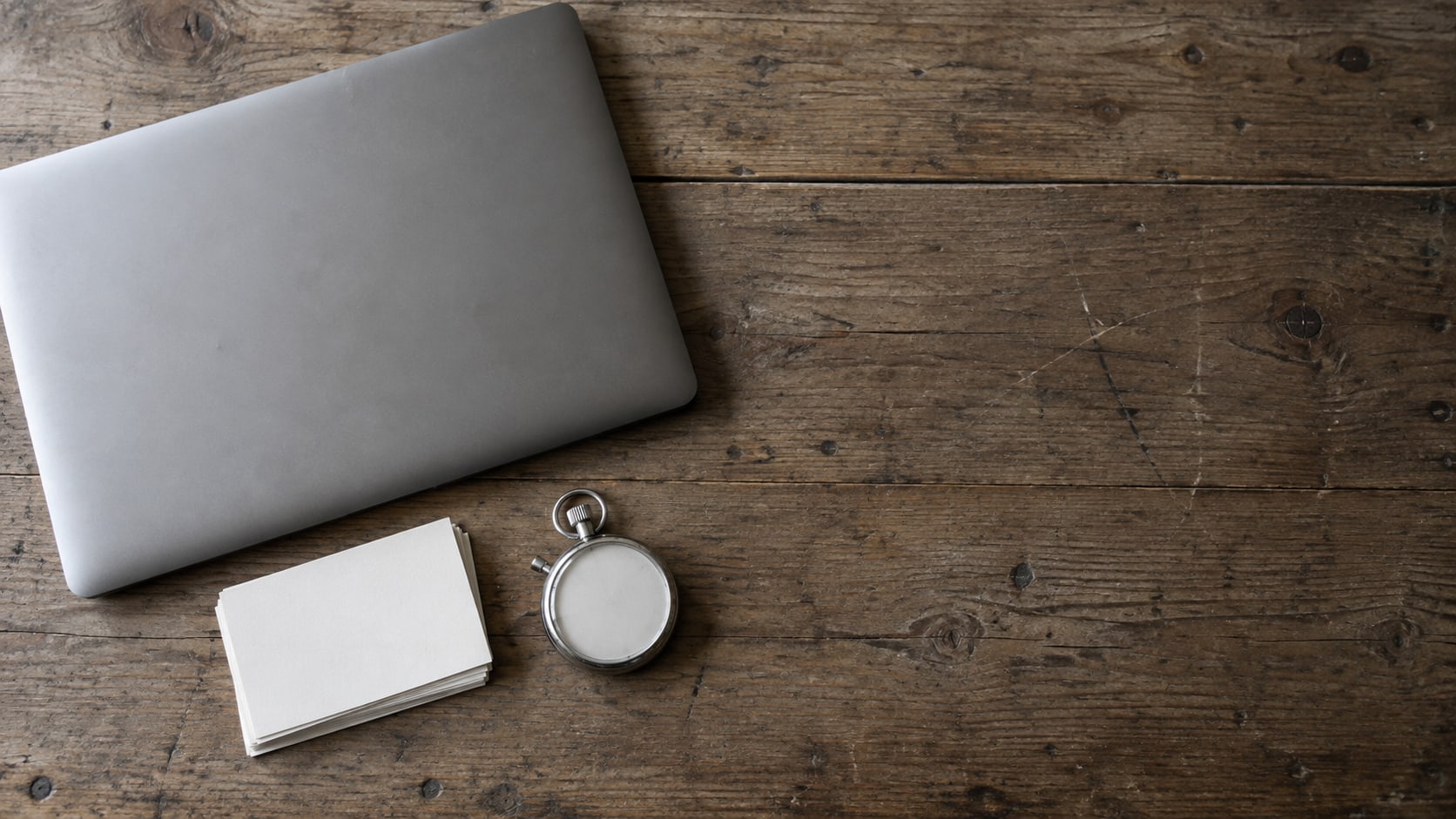 A closed laptop, a small stack of blank index cards and a stopwatch on a worn wooden desk, photographed from above.