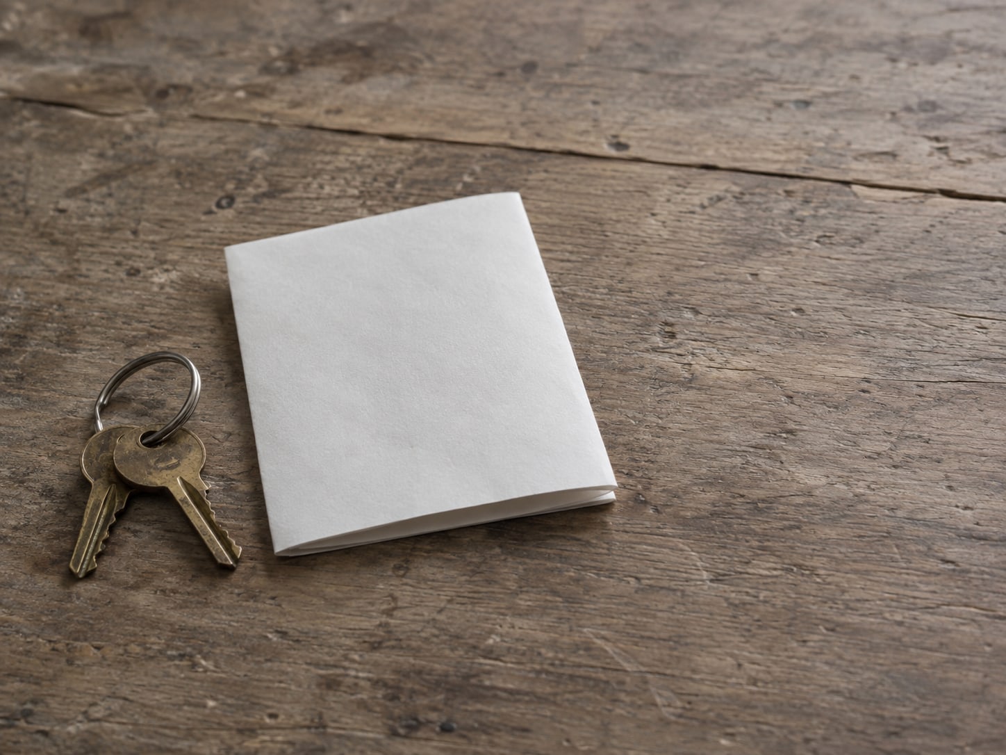 Two small keys on a plain ring next to a folded blank sheet of paper on a wooden table.