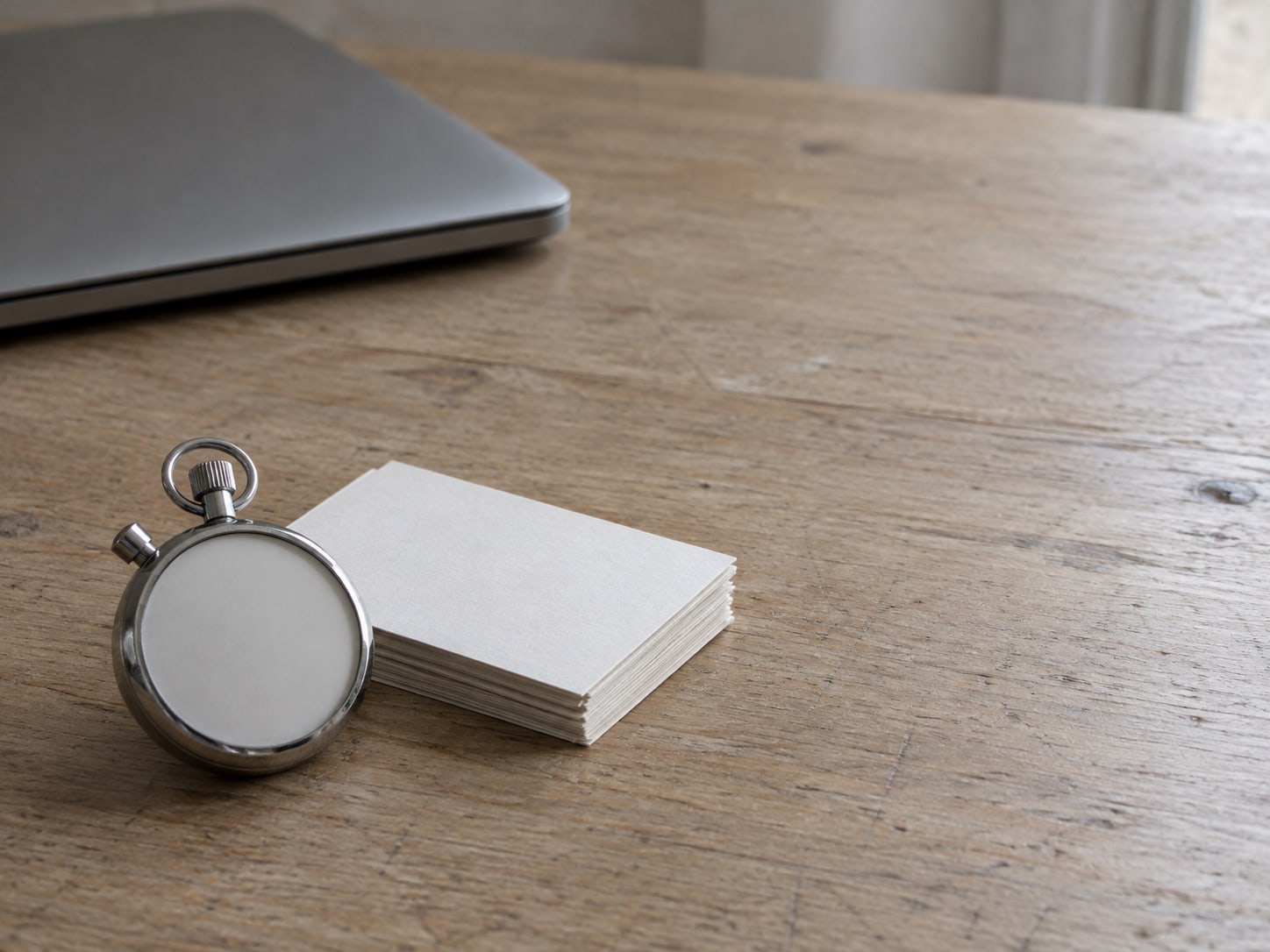 A stopwatch and a small stack of index cards on a plain wooden desk beside a closed laptop.