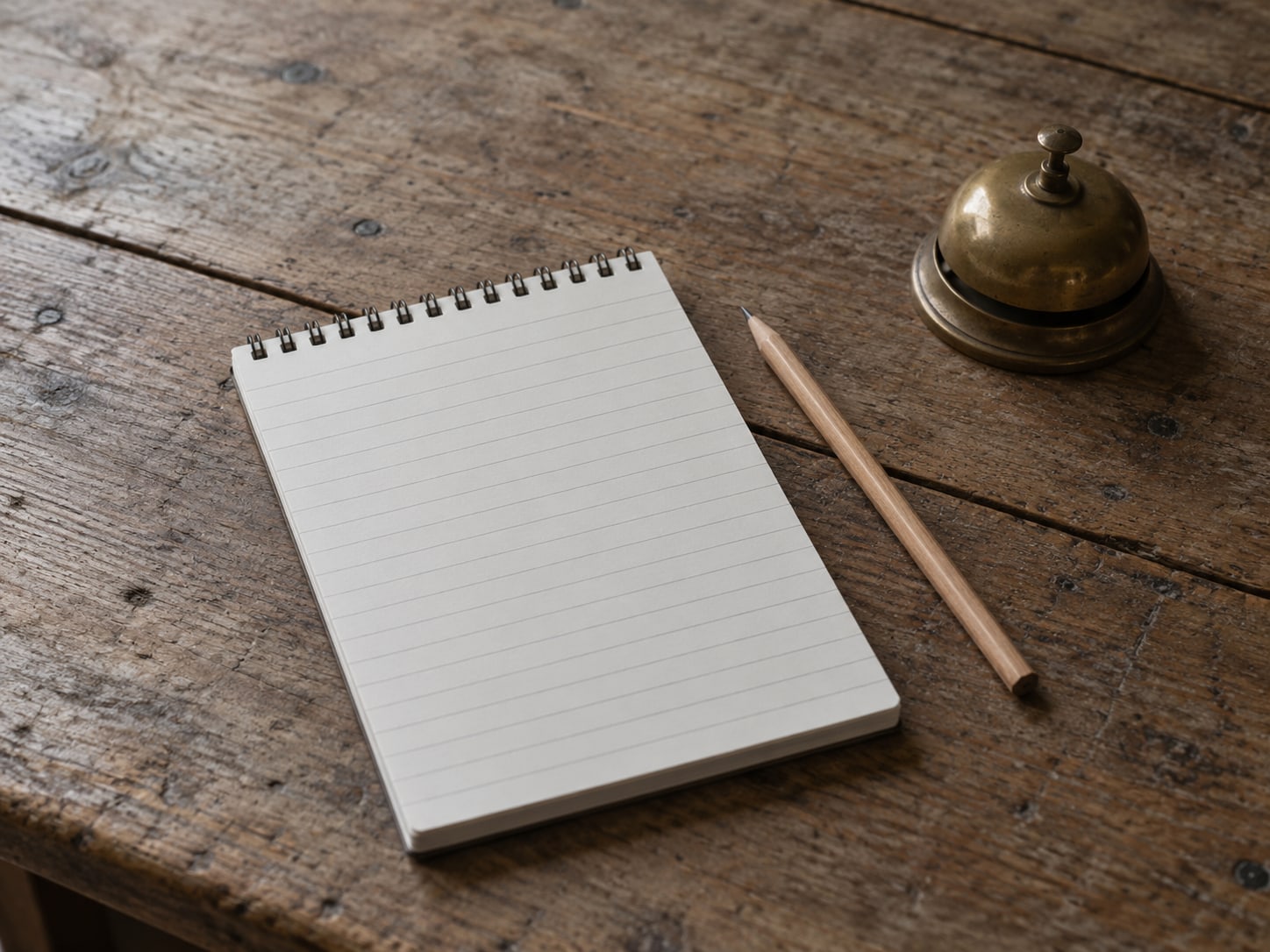 A single ruled notepad with blank lines beside a pencil and a small brass bell on a wooden table.
