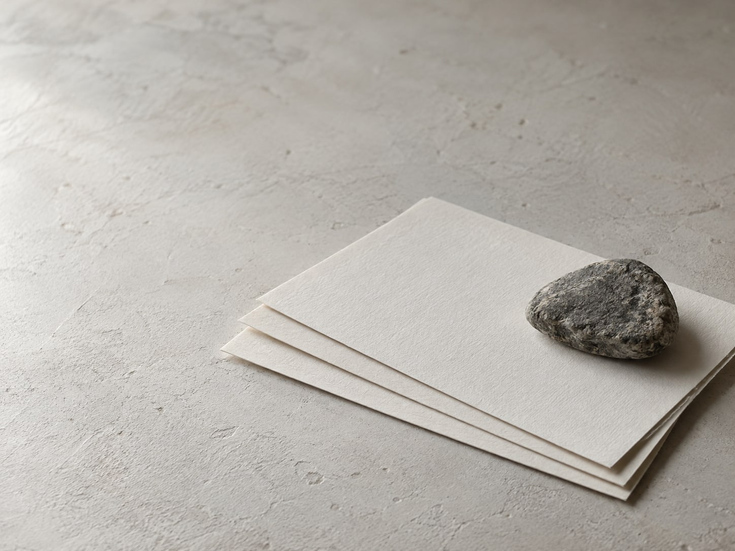 A neat stack of three blank sheets of paper held under a small stone on a clean desk.