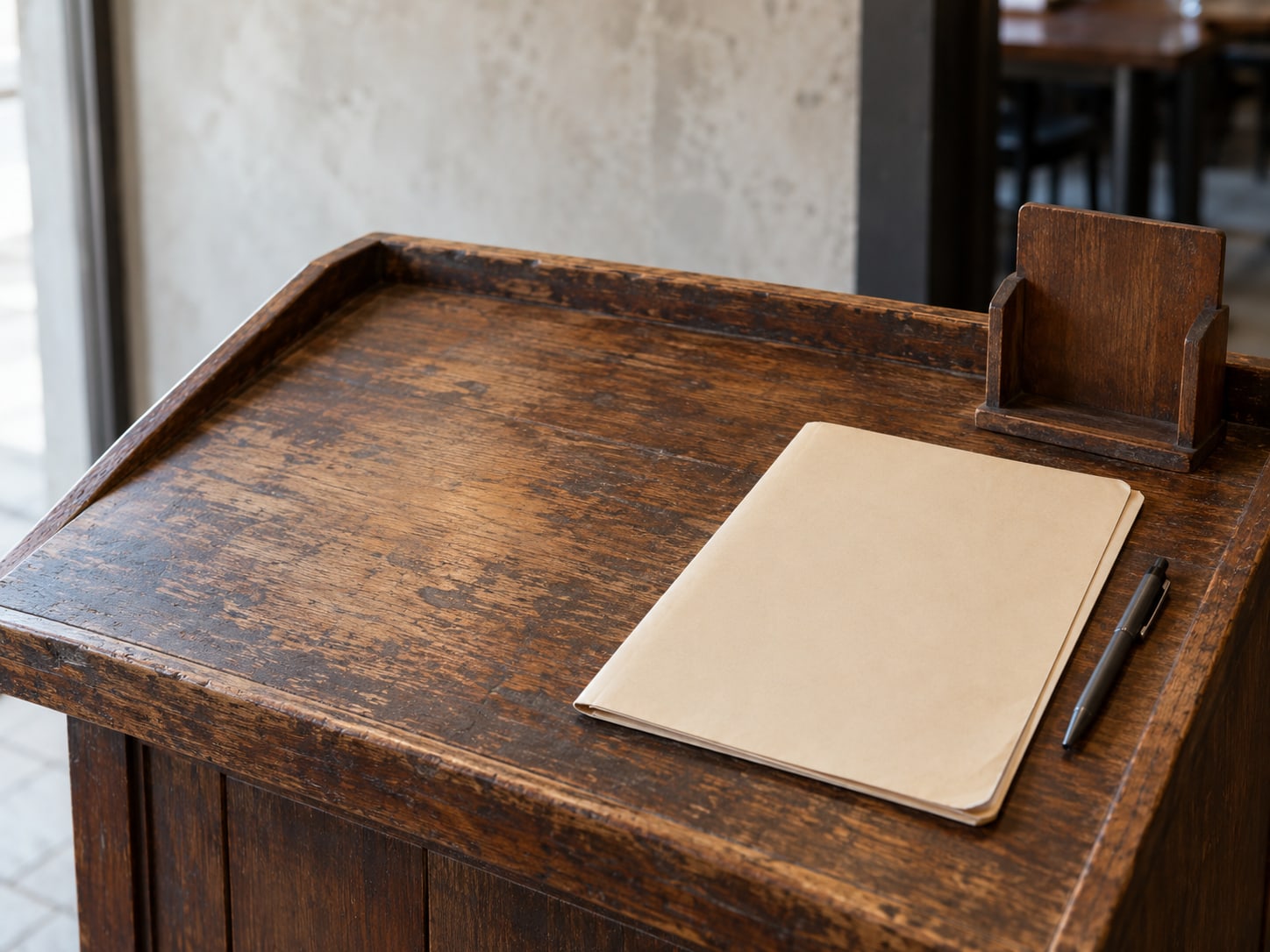 A closed manila folder and a pen resting on a restaurant host stand beside a small empty menu holder.