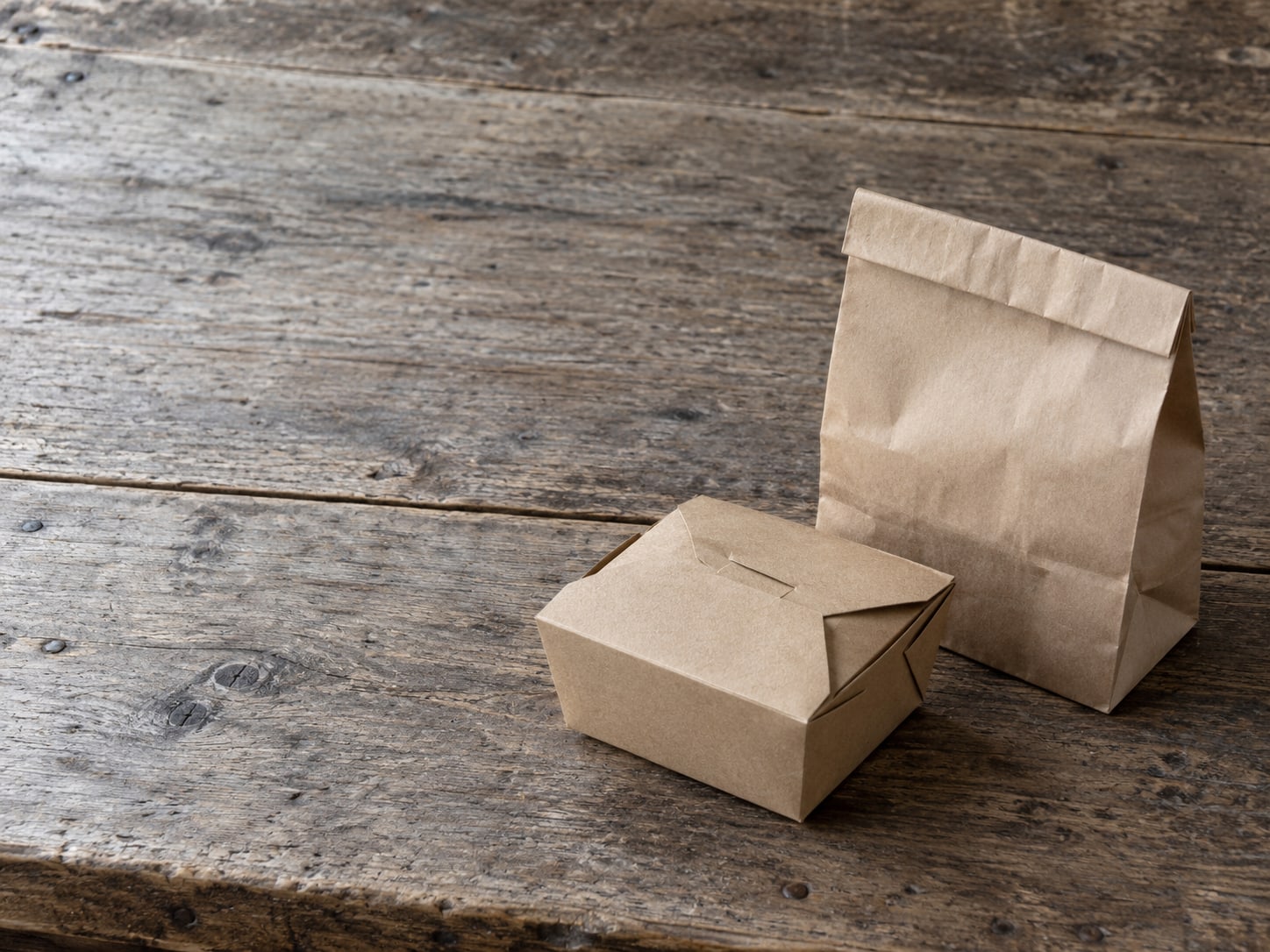 An empty takeout box beside a folded paper bag on a wooden table.