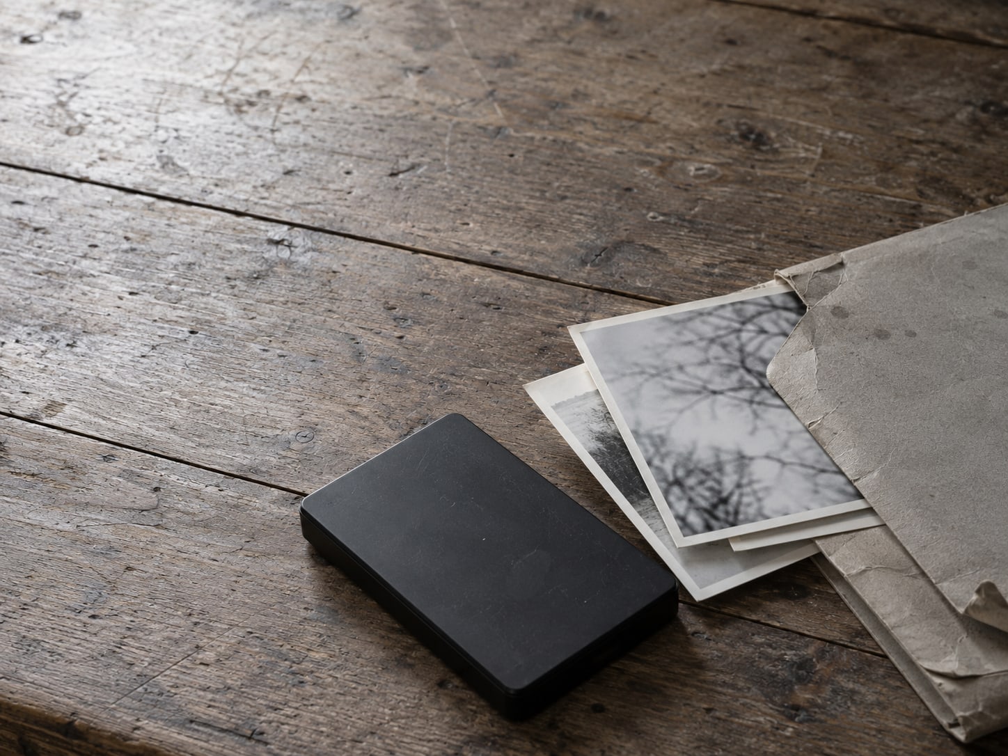 Un disco duro externo negro y una carpeta de fotos impresas sobre una mesa gastada.