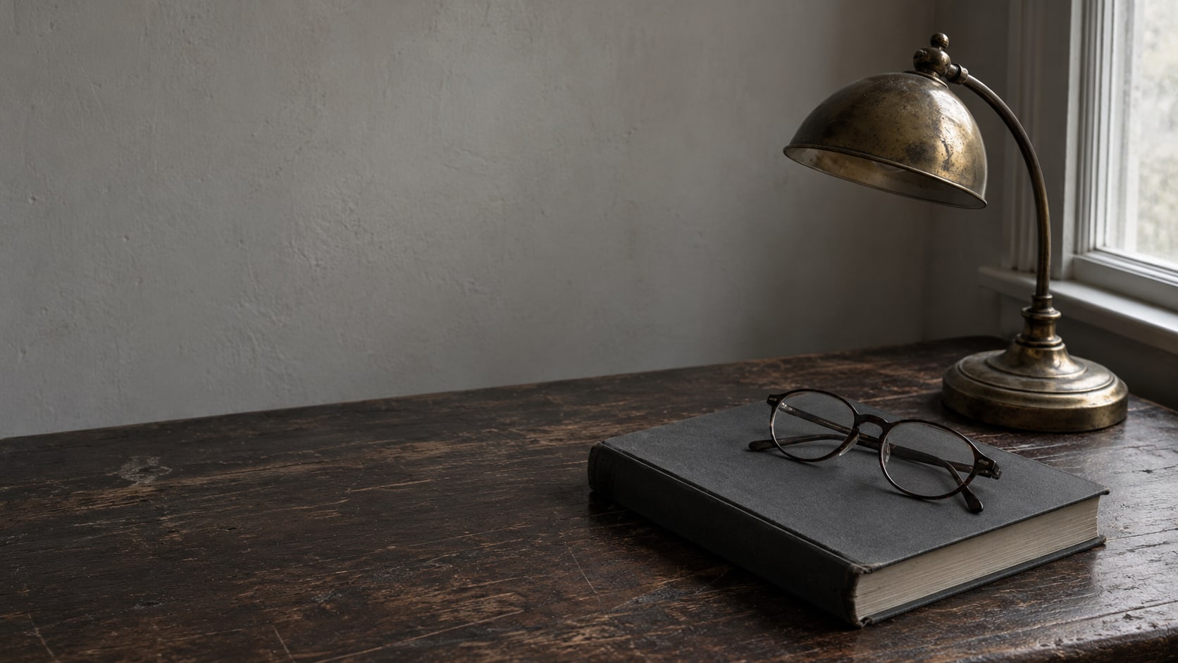 Un par de lentes de lectura apoyados sobre un libro cerrado de tapa dura, junto a una pequeña lámpara de latón sobre un escritorio de madera oscura.