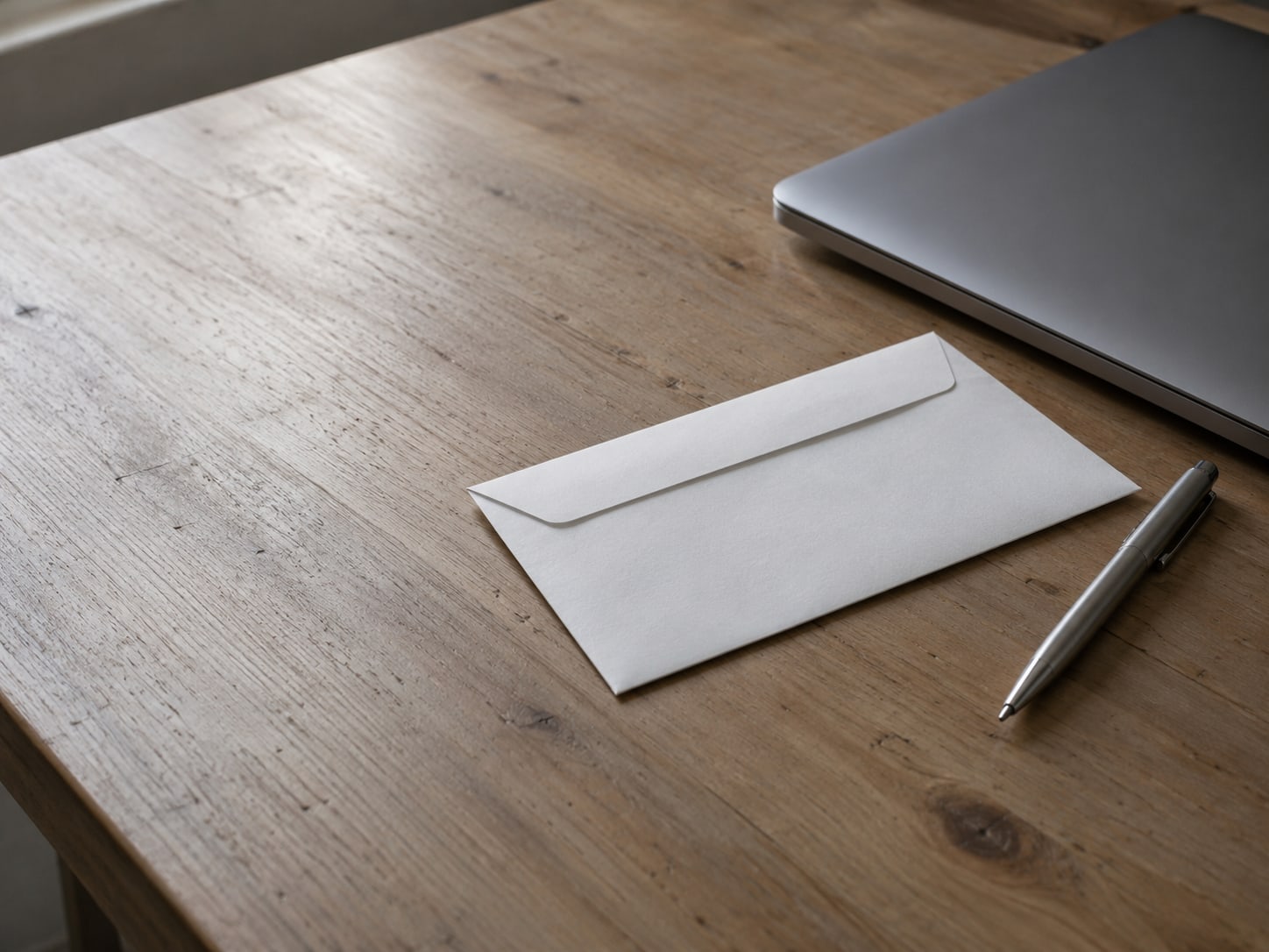 A sealed blank envelope and a pen resting beside a closed laptop on a tidy wooden desk.