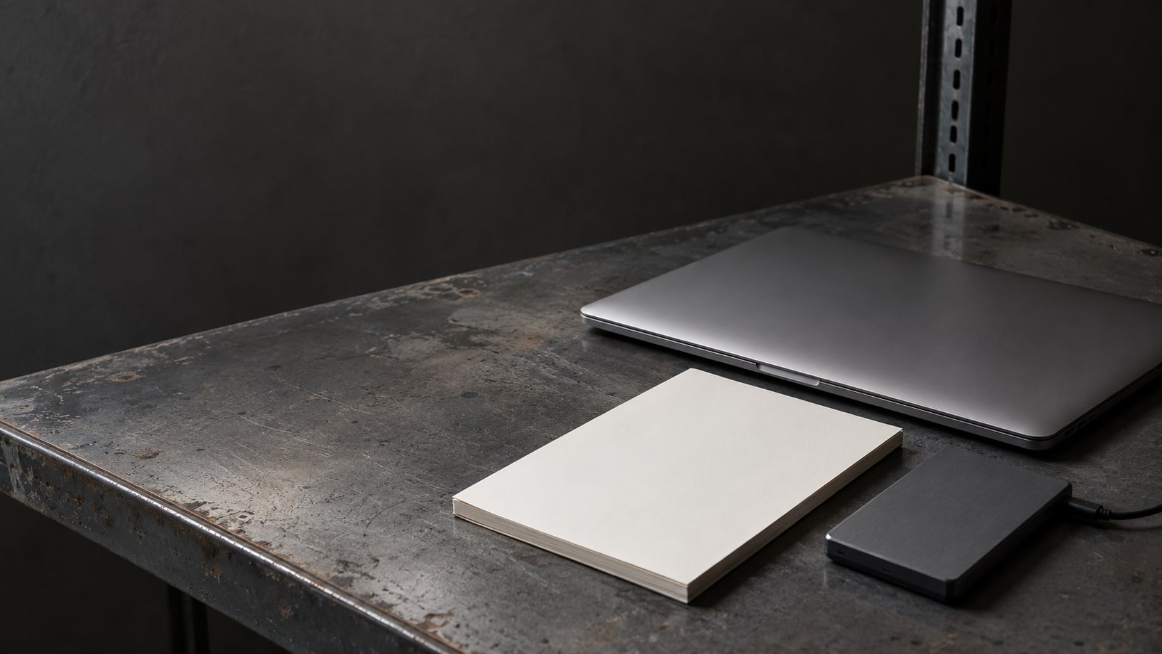 A closed laptop, a blank notepad and a small external drive arranged neatly on a server rack shelf.