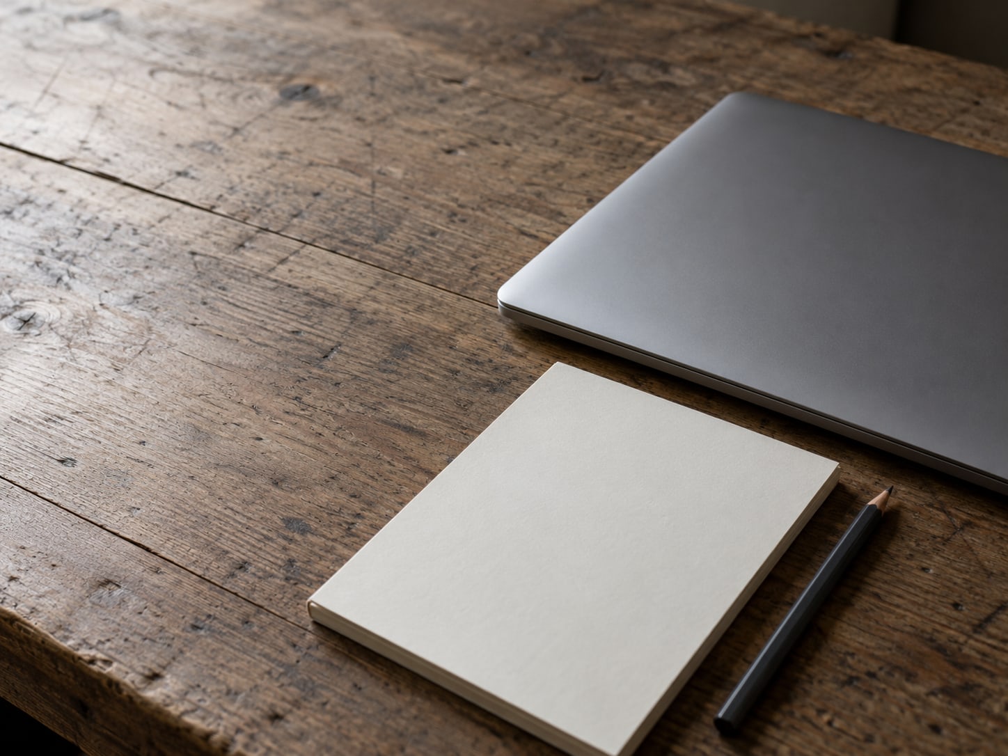 A closed laptop beside a blank notepad and a sharpened pencil on a worn wooden desk.