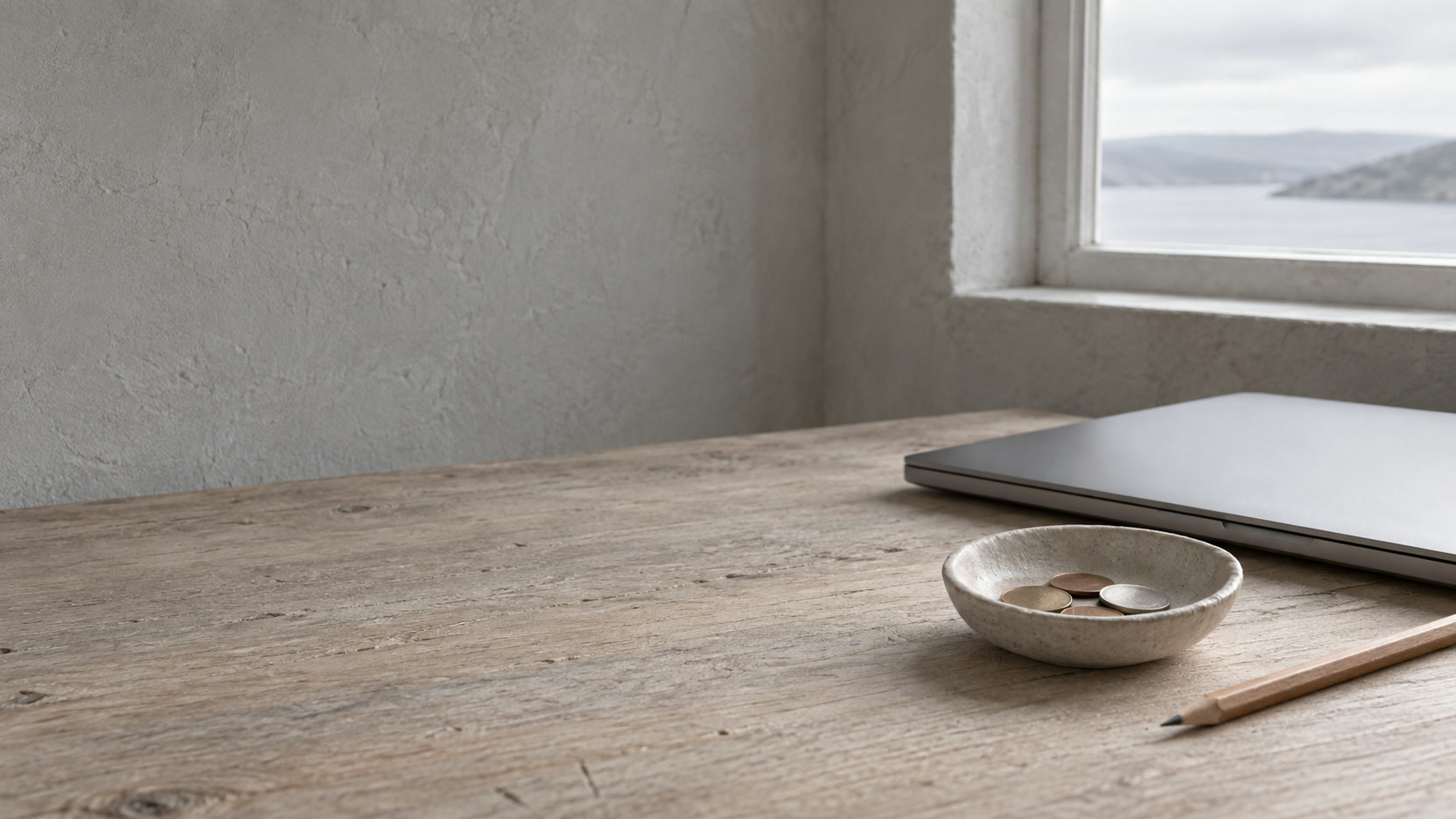 A small ceramic dish holding a few coins beside a closed laptop and a pencil on a pale oak desk.
