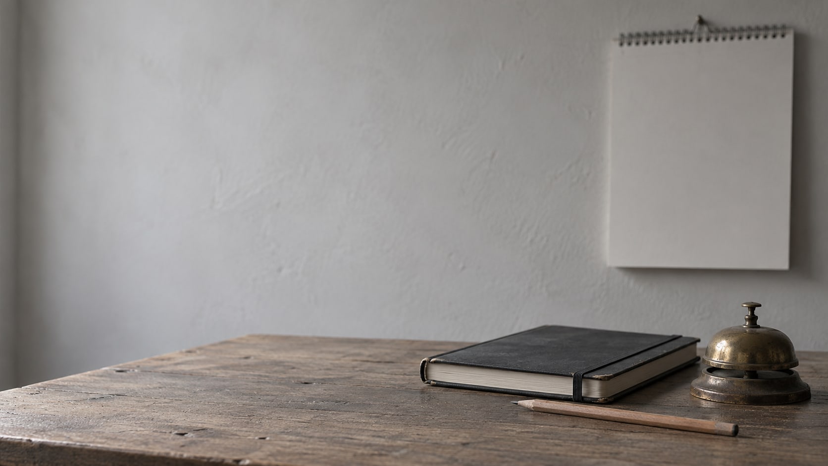 A closed appointment book, a pencil and a small brass desk bell on a wooden counter, with a blank wall calendar softly out of focus behind them.