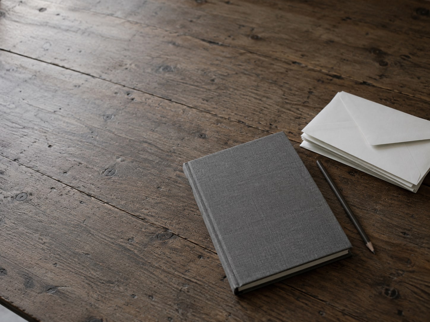 Un cuaderno cerrado y un lápiz junto a una pila de sobres sin abrir sobre un escritorio de madera gastado.