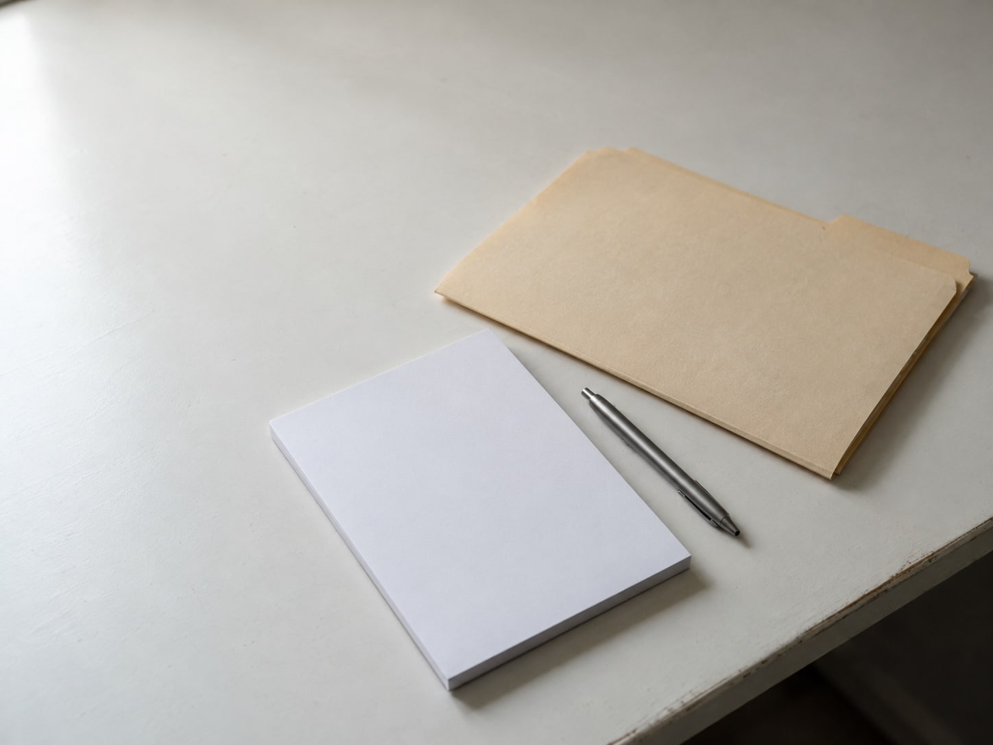 A closed manila folder and a pen lying beside a blank notepad on a clean desk.