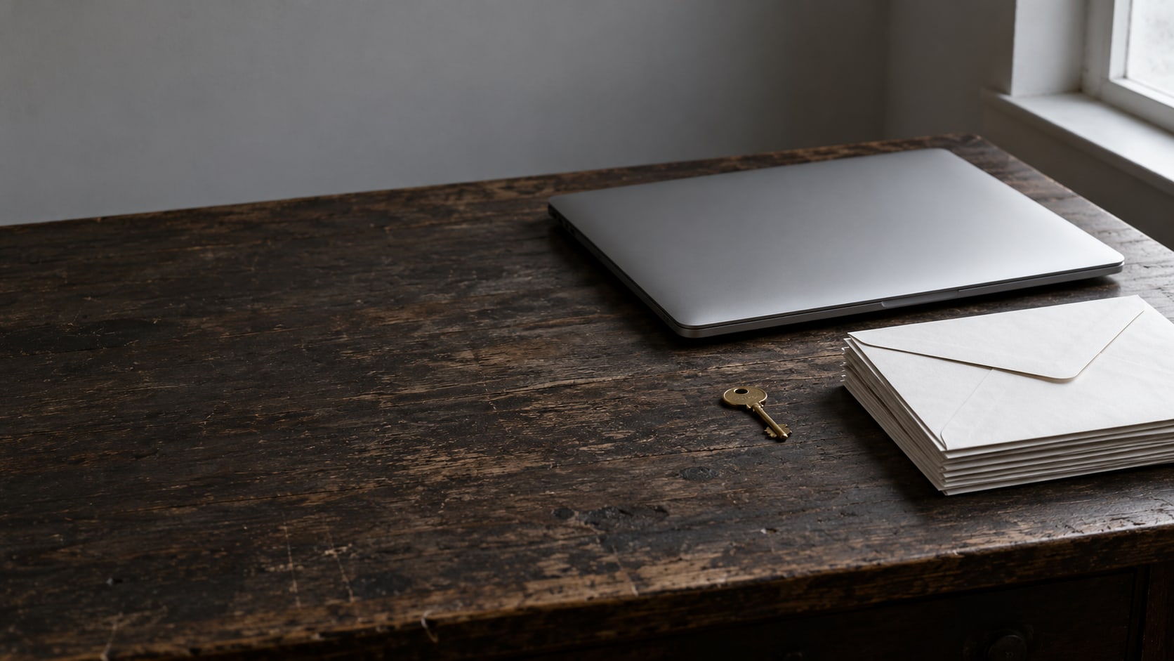 A closed laptop beside a small brass key and a stack of sealed envelopes on a dark wooden desk.