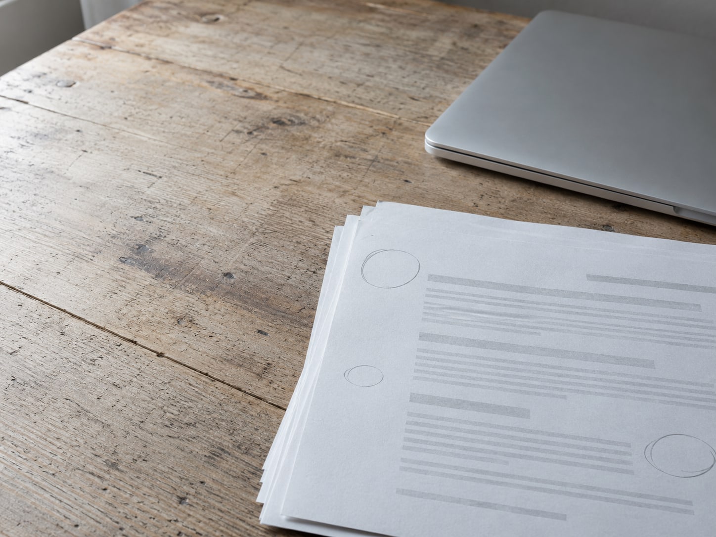 A small stack of printed report pages with a few pencil circles in the margin, beside a closed laptop on a wooden desk.