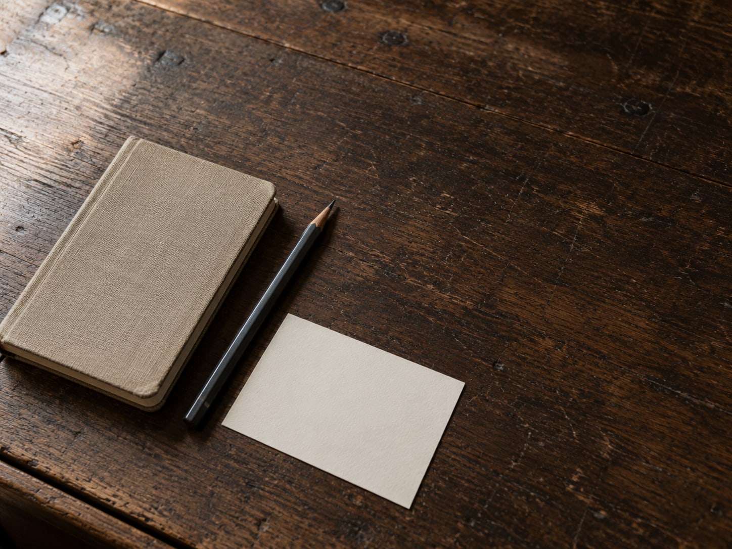 A closed notebook beside a pencil and a single unmarked index card on a worn wooden desk.
