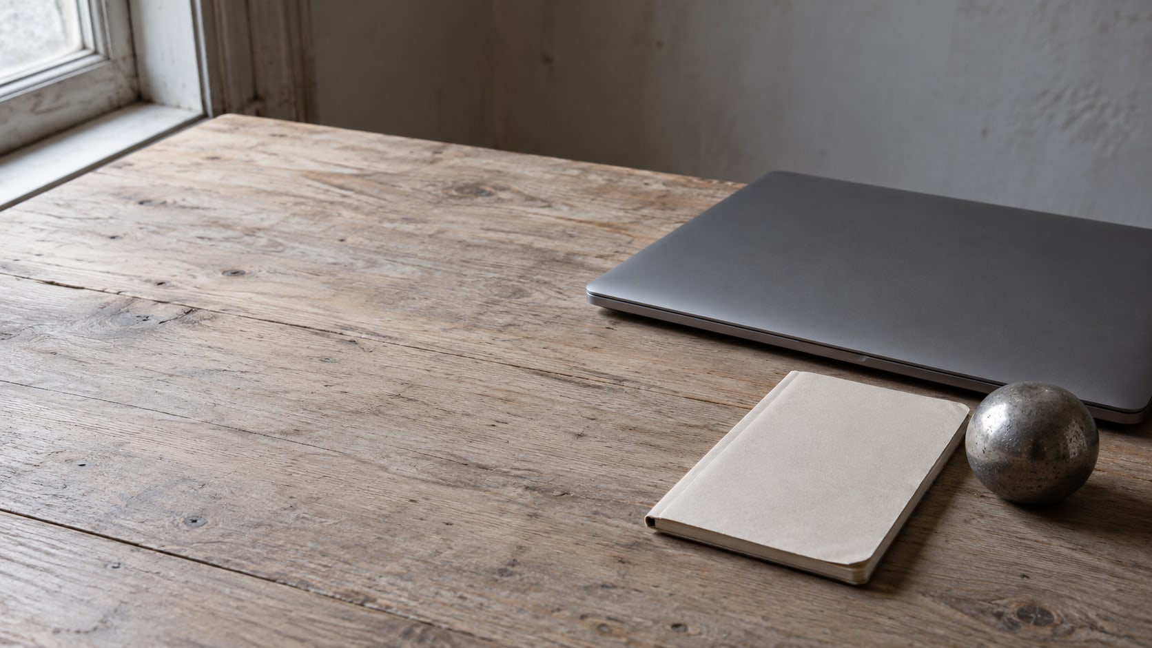 A closed laptop, a small notebook and a paperweight arranged on a clean wooden desk in soft morning light.