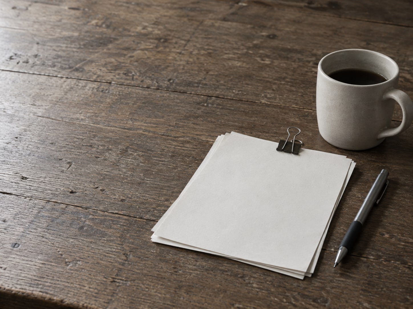 A few sheets of blank paper held by a binder clip next to a pen and a coffee mug on a wooden table.