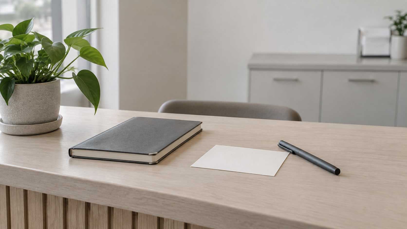 A closed appointment book, a blank notecard and a capped pen on a quiet clinic reception counter beside a small potted plant.