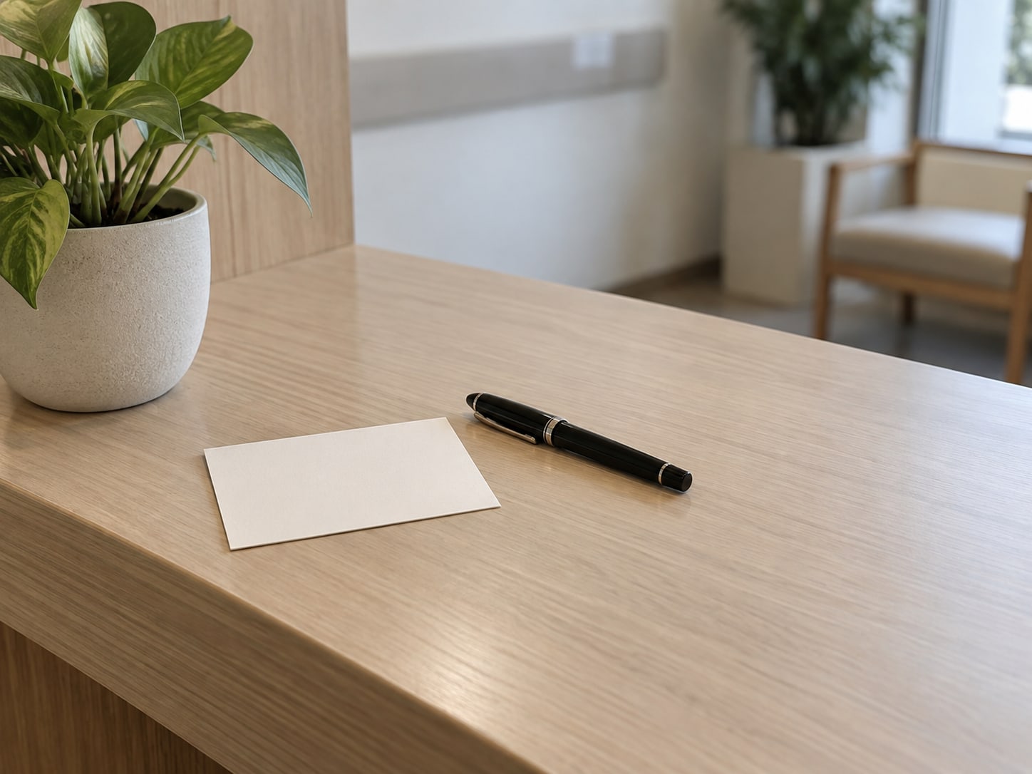 A blank notecard and a closed fountain pen resting on a clean wooden reception counter beside a small potted plant.