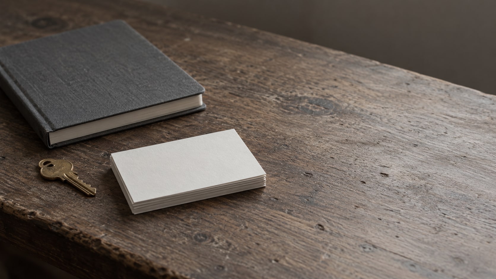 Una llave de latón en forma de casa y un cuaderno cerrado sobre un escritorio de madera, junto a una pequeña pila de tarjetas en blanco.