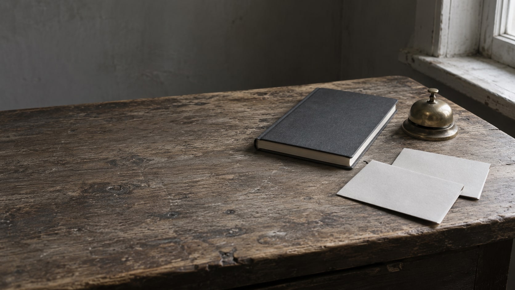 A closed notebook, a small brass desk bell and two blank folded papers set side by side on a wooden desk.