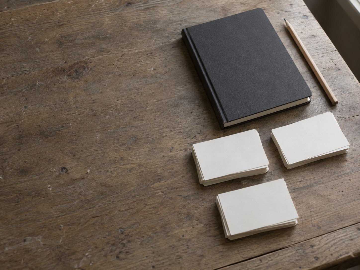 A closed notebook and a pencil beside a small stack of blank index cards sorted into three separate piles on a wooden desk.