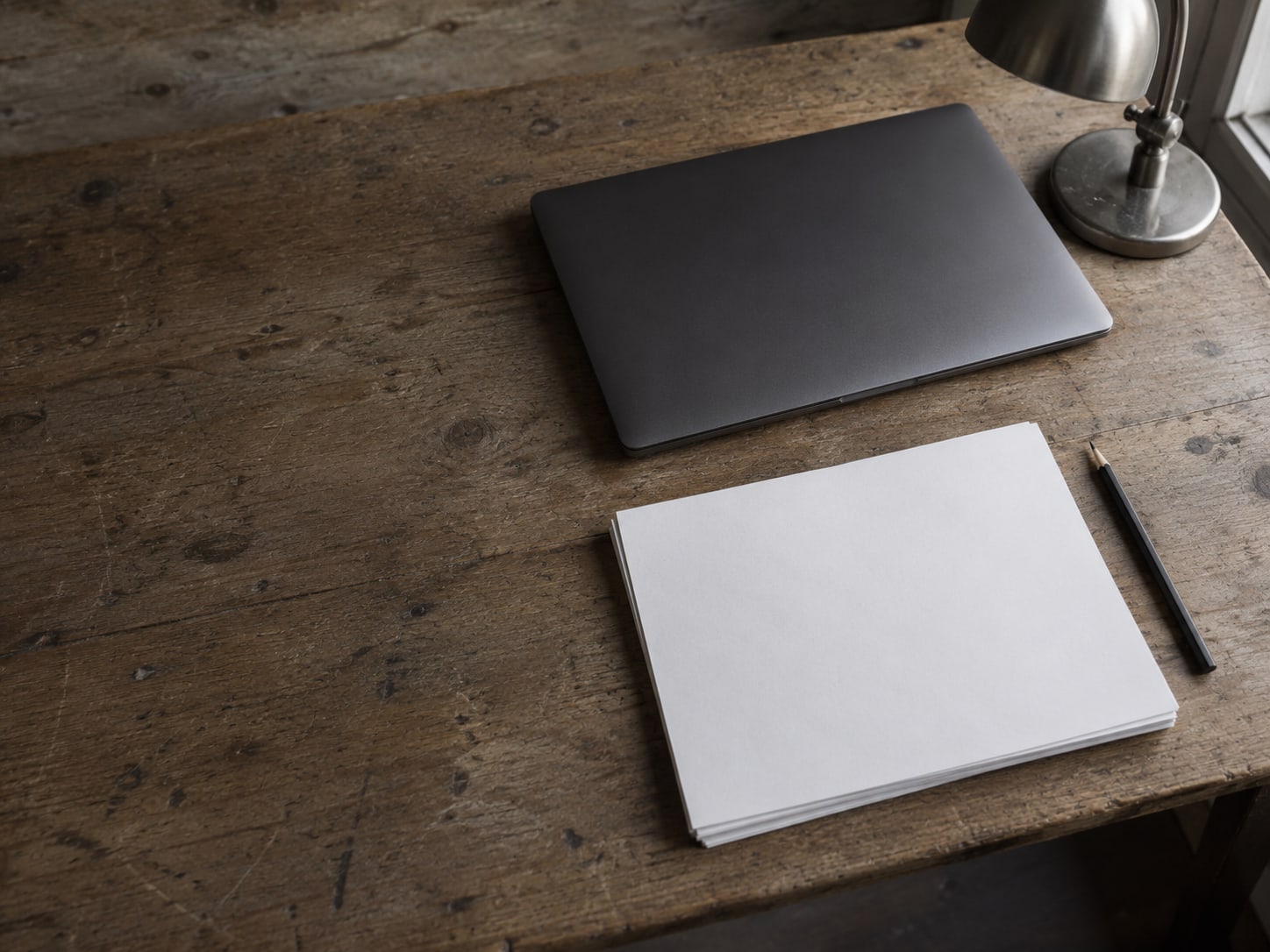 A closed laptop beside a stack of blank paper and a pencil on a worn wooden desk, under a small desk lamp.