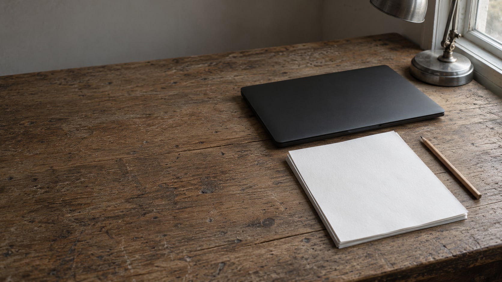 A closed laptop and a small stack of blank papers beside a pencil on a worn wooden desk under a lamp.