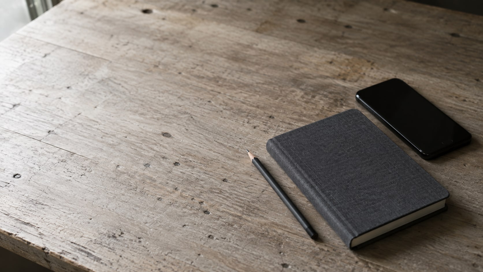 Un cuaderno cerrado, un lápiz y un teléfono apagado boca abajo sobre un mostrador de madera liso.