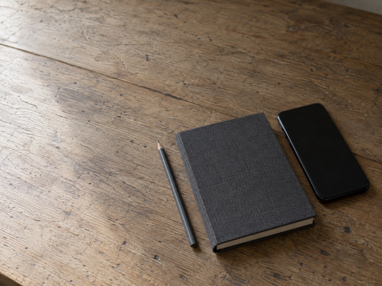 Un cuaderno cerrado y un lápiz junto a un teléfono apagado boca abajo sobre un mostrador de madera.