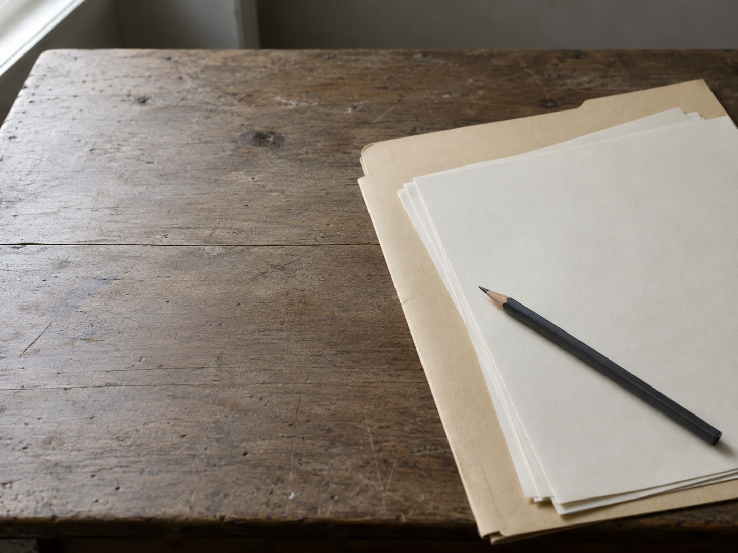 Una carpeta manila con algunas hojas sueltas en blanco y un lápiz sobre un escritorio de madera gastada.