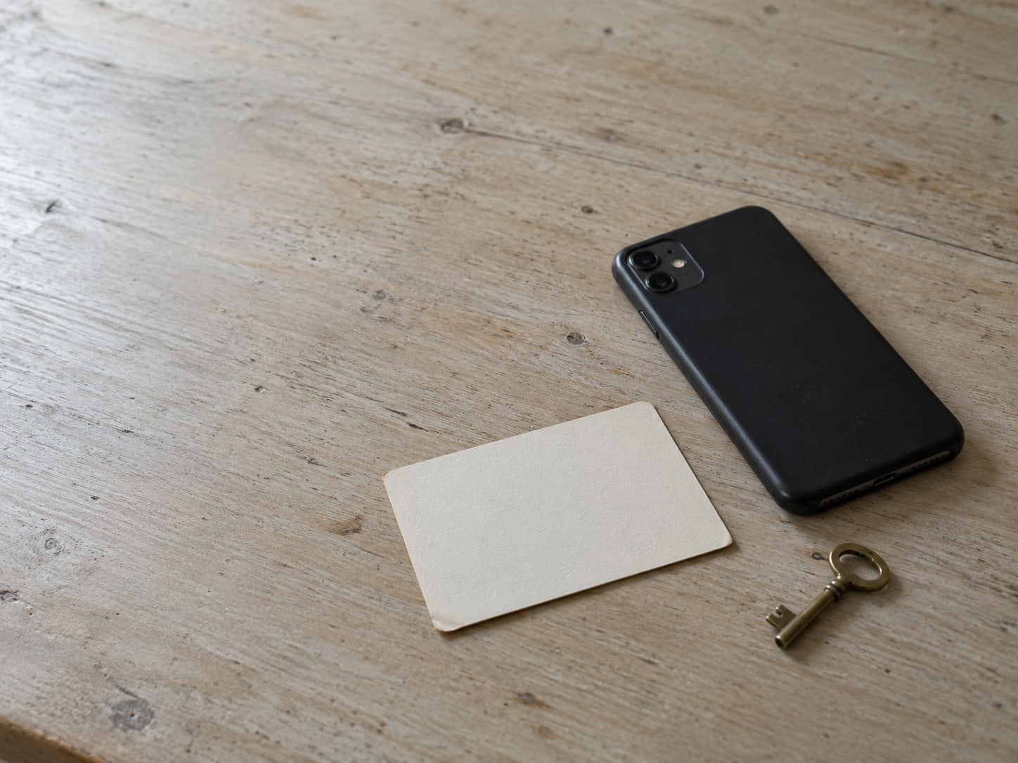 A phone lying face down on a wooden desk beside a blank index card and a small brass key.