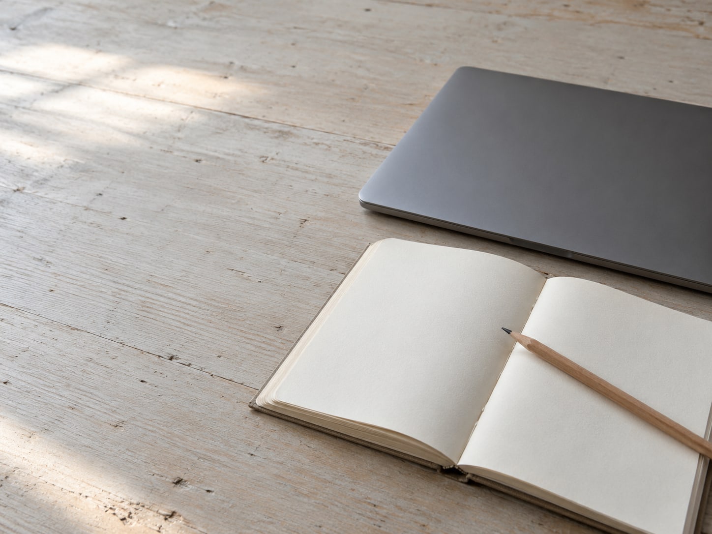 A closed laptop beside a notebook with blank pages and a pencil resting across it, on a pale wooden desk.