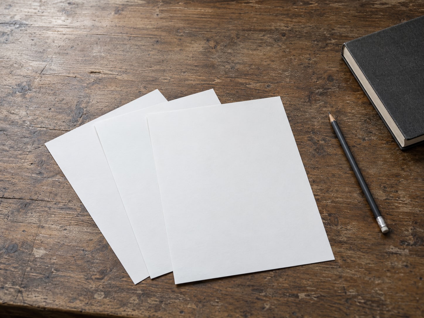 Three blank sheets of paper fanned out on a worn wooden desk beside a pencil and a closed notebook.