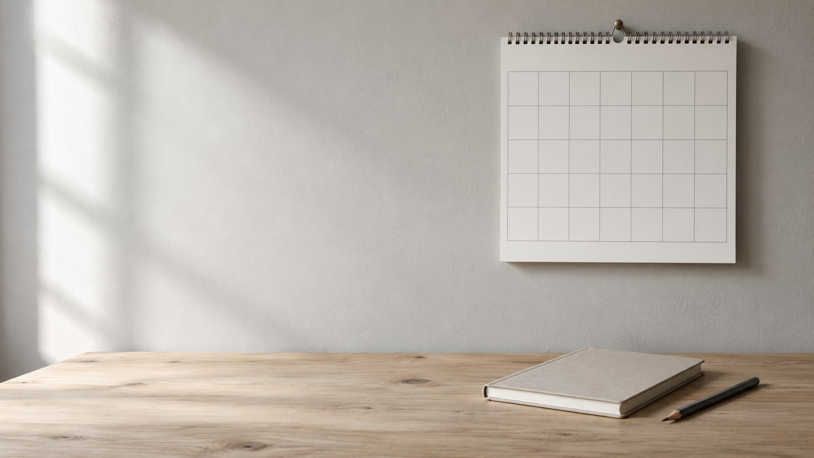 A wall calendar, a closed notebook and a pencil arranged on a light wooden desk.