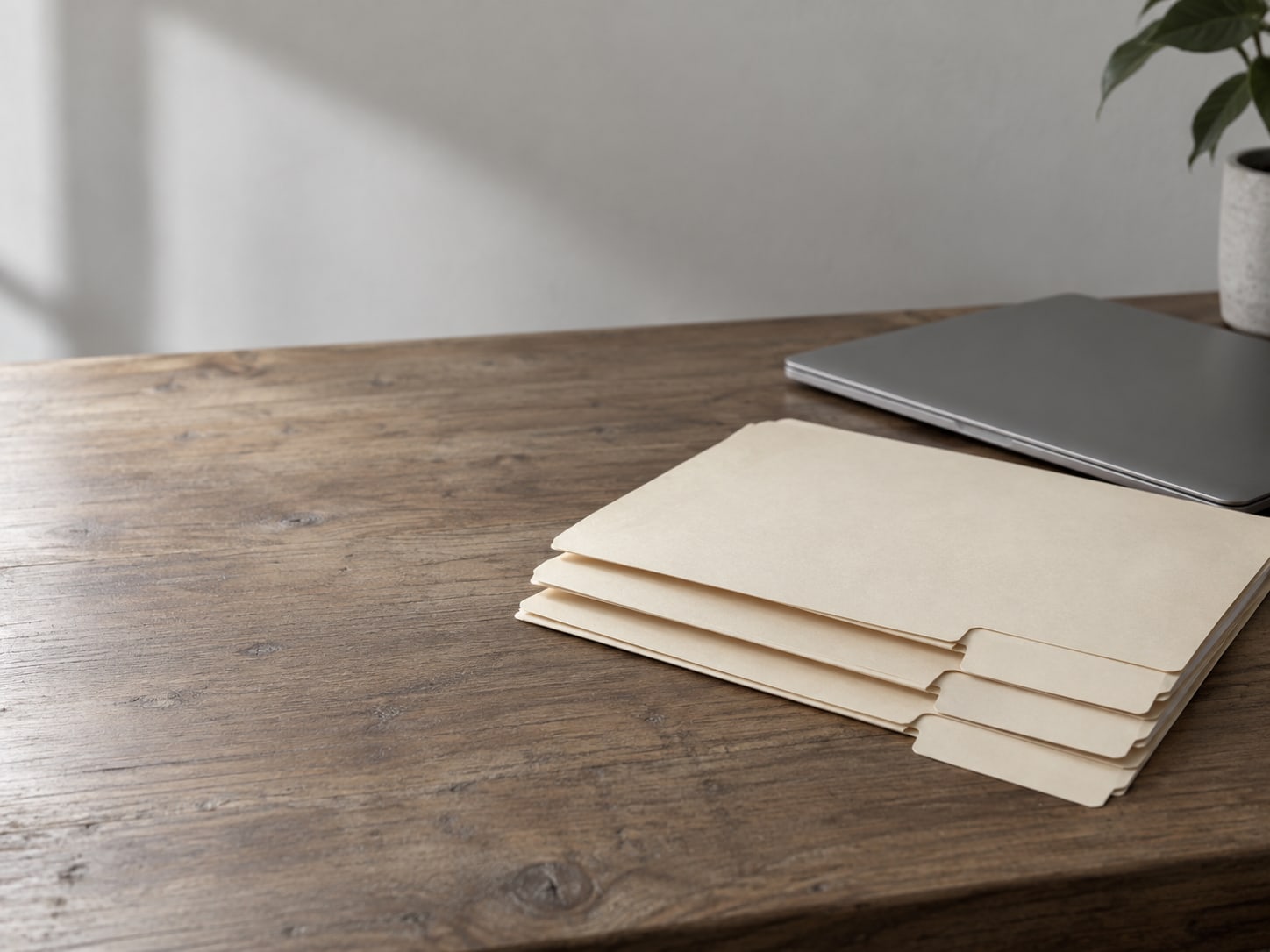 Three plain manila folders with blank tabs stacked neatly on a wooden desk beside a closed laptop.