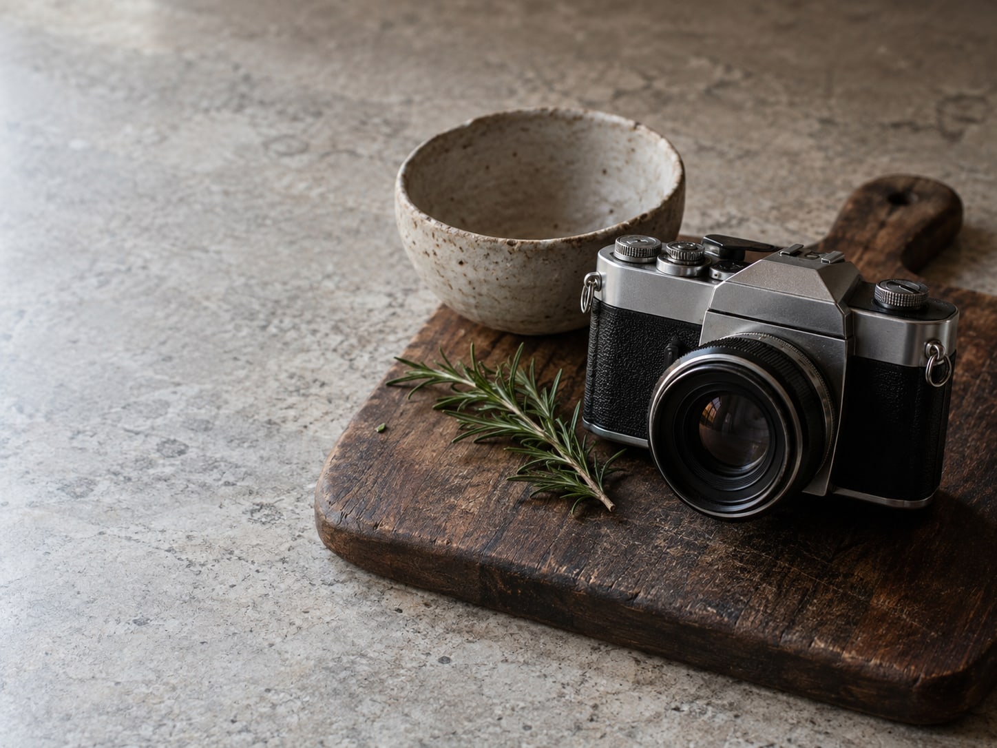 A camera resting on a walnut cutting board beside a hand-thrown bowl and a sprig of rosemary on a kitchen counter.