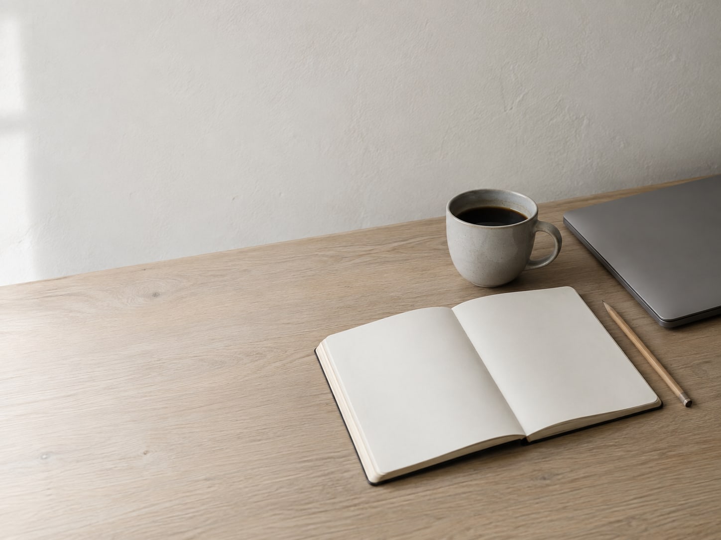 A notebook open to a blank page on a desk beside a closed laptop, a pencil and a cup of coffee.