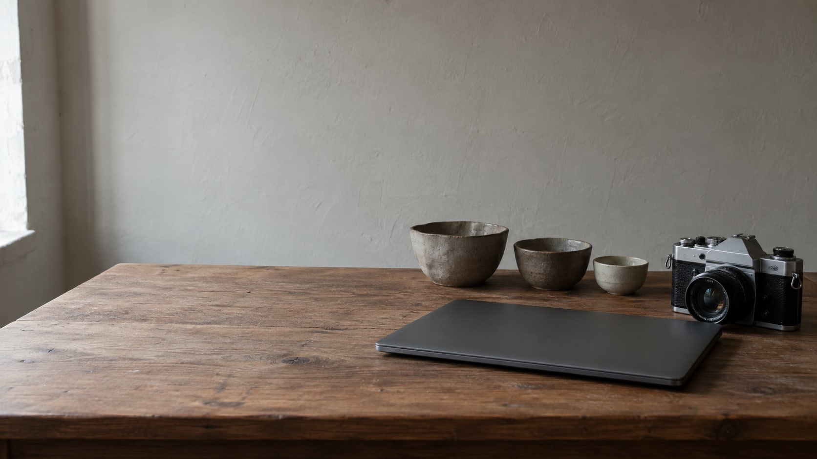 A closed laptop and a camera resting on a wooden table beside a small row of handmade ceramic bowls.