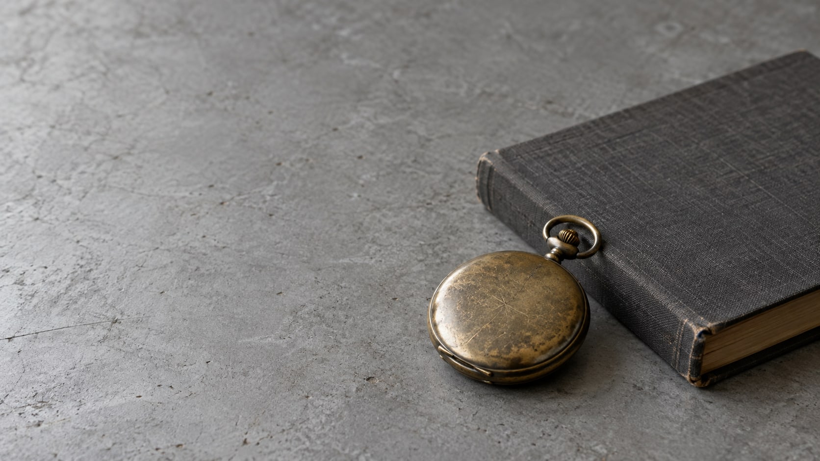 A brass pocket watch lying beside a closed ledger on a plain gray table.