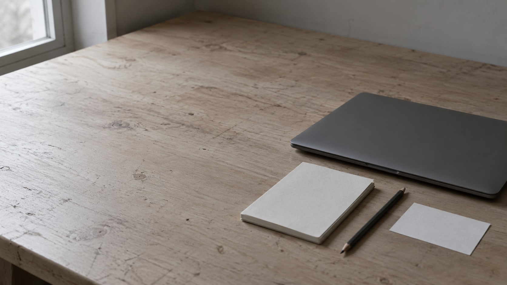 Una laptop cerrada, una libreta pequeña y un lápiz junto a una ficha en blanco sobre un escritorio de madera limpio.