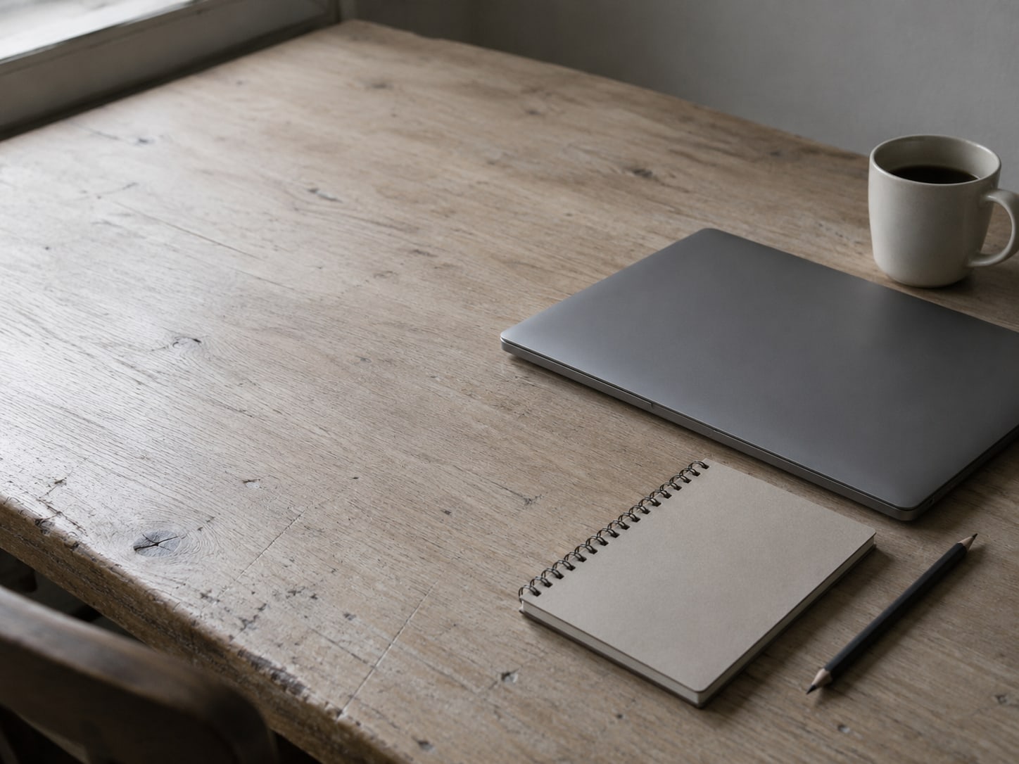 Una laptop cerrada, un cuaderno de espiral y un lápiz sobre un escritorio de madera sencillo, junto a una taza.