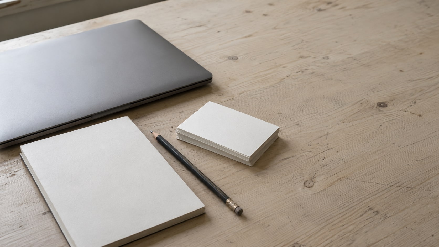 A notepad and pencil beside a closed laptop and a stack of blank index cards on a clean wooden desk.