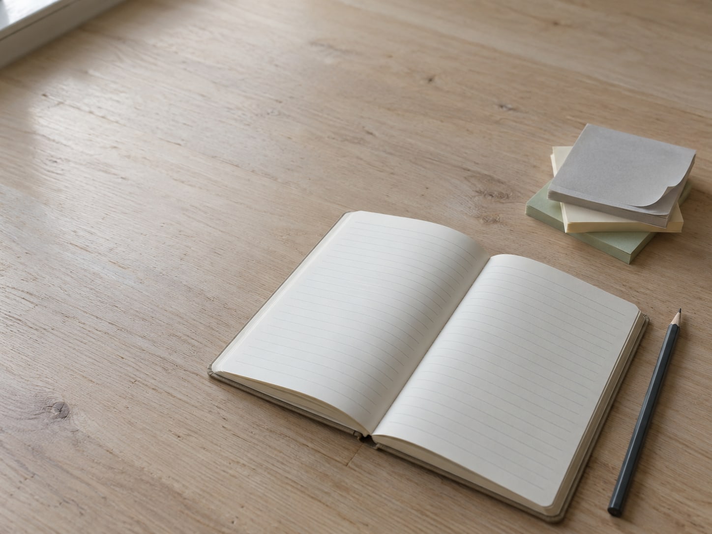 A paper notebook open to blank lined pages beside a pencil and a stack of sticky notes in three colors.
