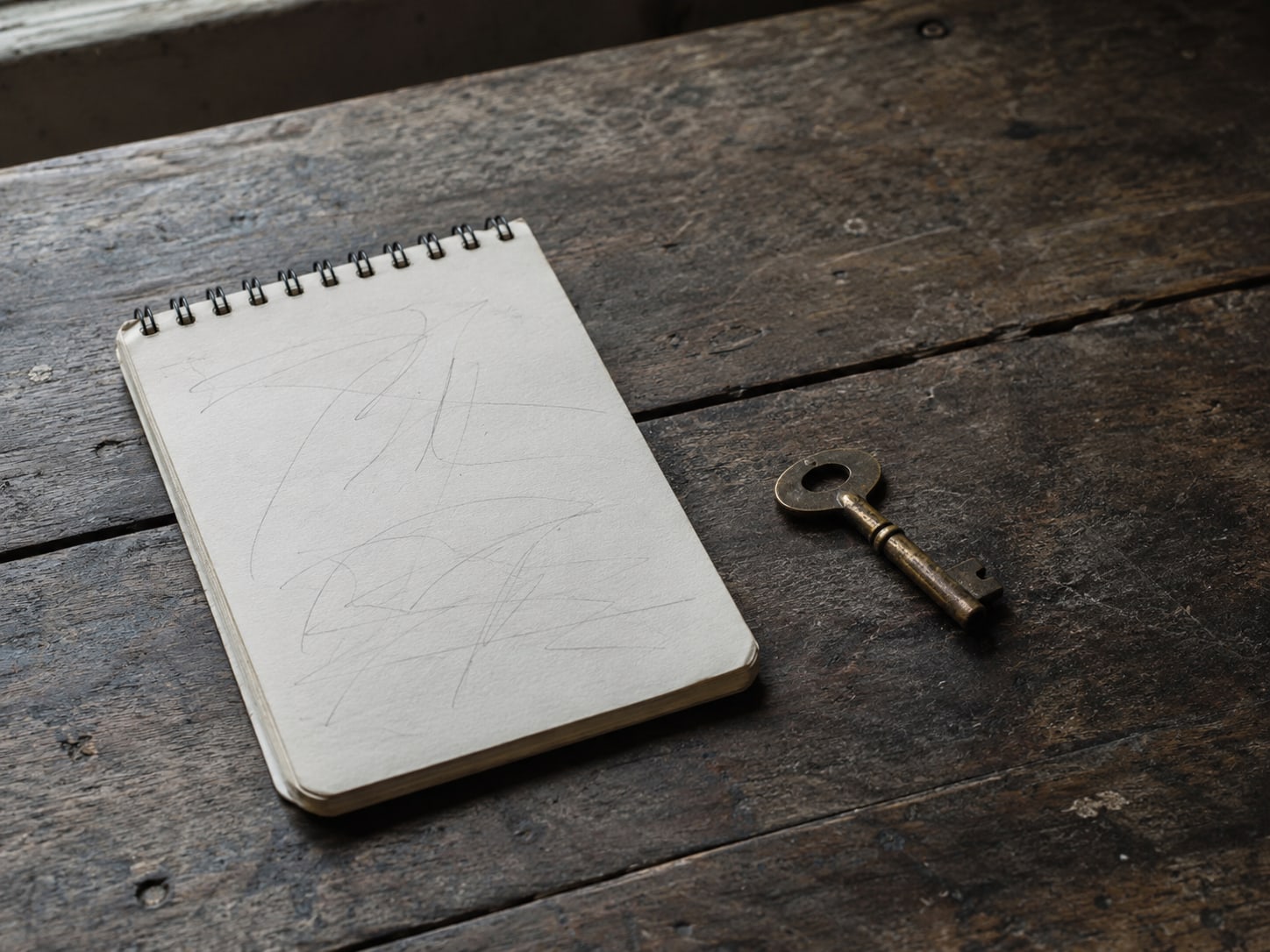 Un bloc de notas liso con líneas de lápiz ilegibles junto a una pequeña llave de latón sobre una mesa de madera desgastada.