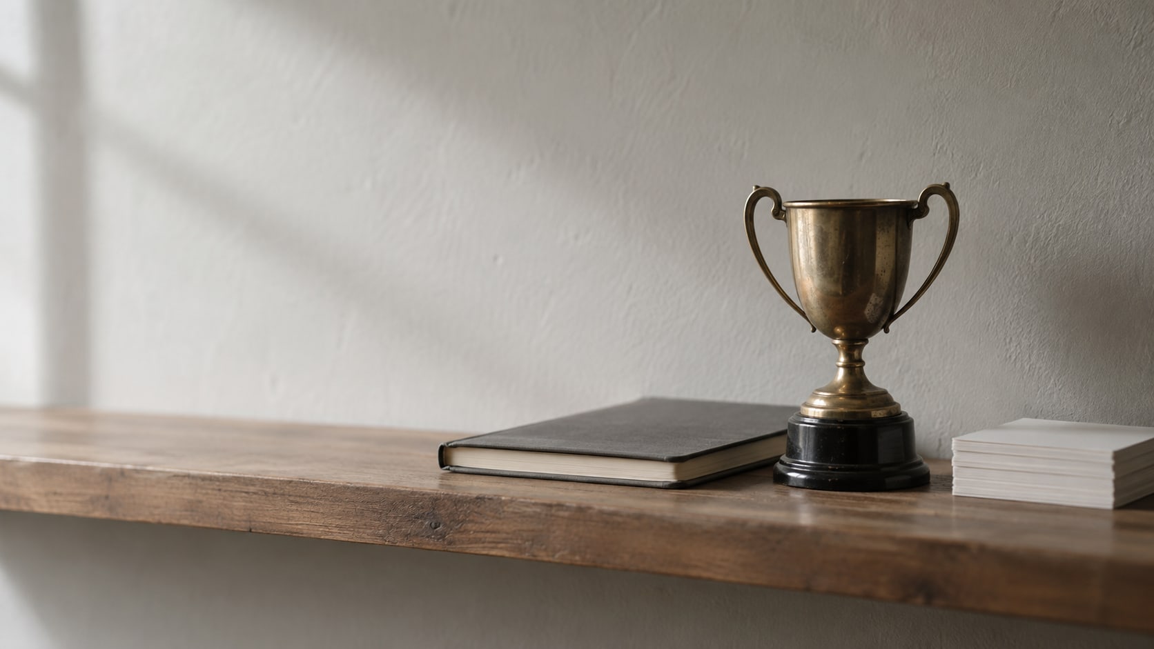 A small brass trophy on a bare wooden shelf beside a closed notebook and a short stack of blank index cards, softly lit from one side.