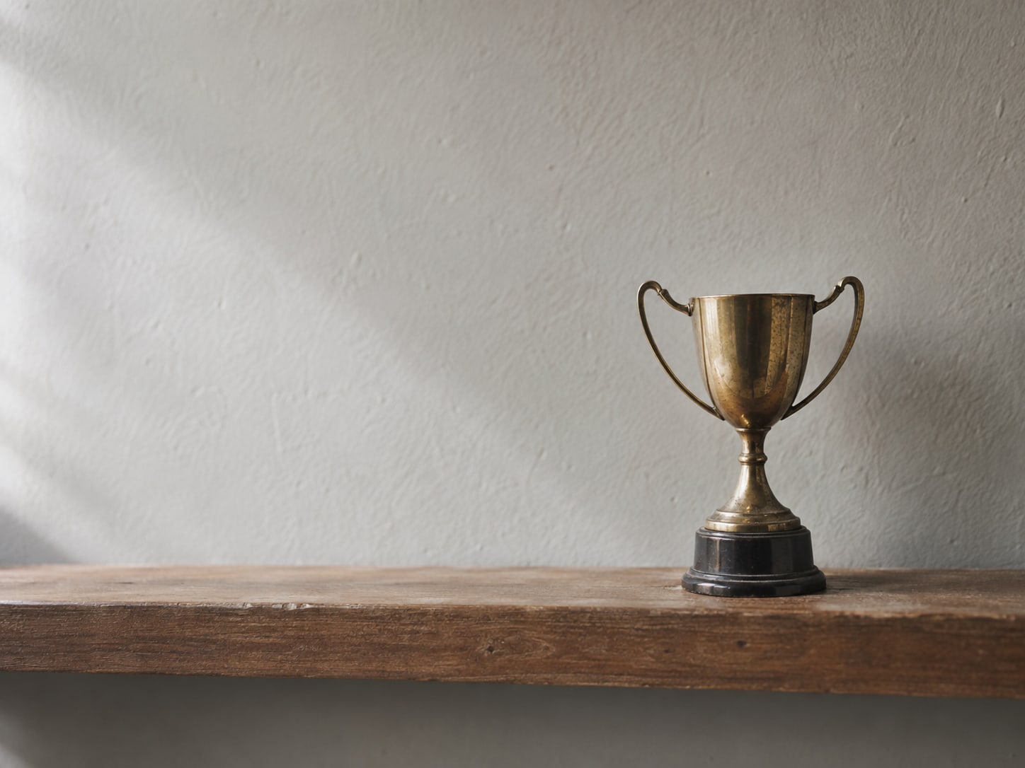 Un solo trofeo pequeño de latón solo sobre un estante de madera desnudo, iluminado suavemente desde un lado.